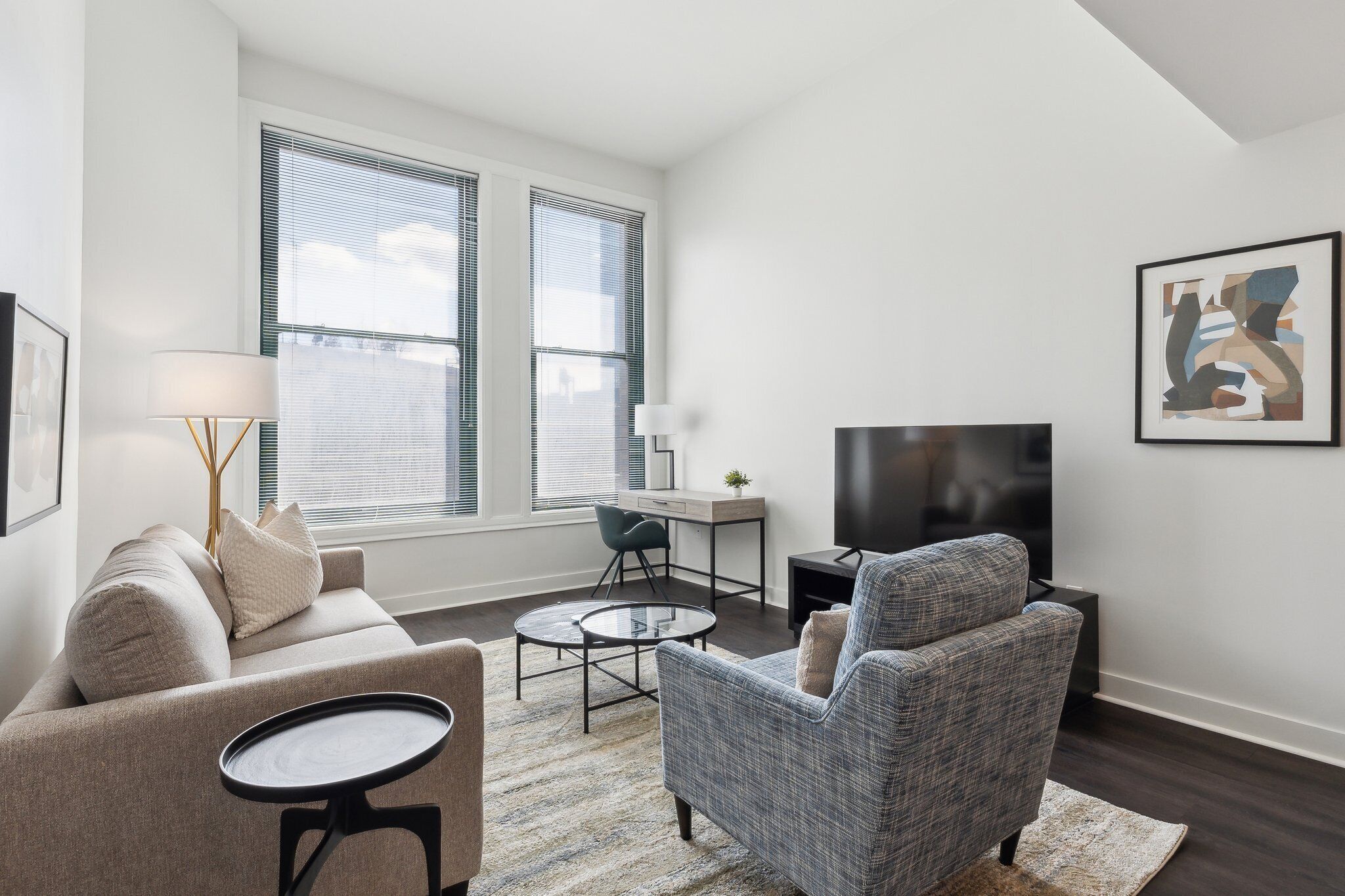 Una sala de estar moderna con sofá, sillón, mesa de centro y televisor. Hay dos ventanas con persianas, un escritorio con silla y un cuadro en la pared.