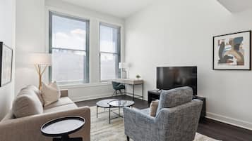 Una sala de estar moderna con sofá, sillón, mesa de centro y televisor. Hay dos ventanas con persianas, un escritorio con silla y un cuadro en la pared.