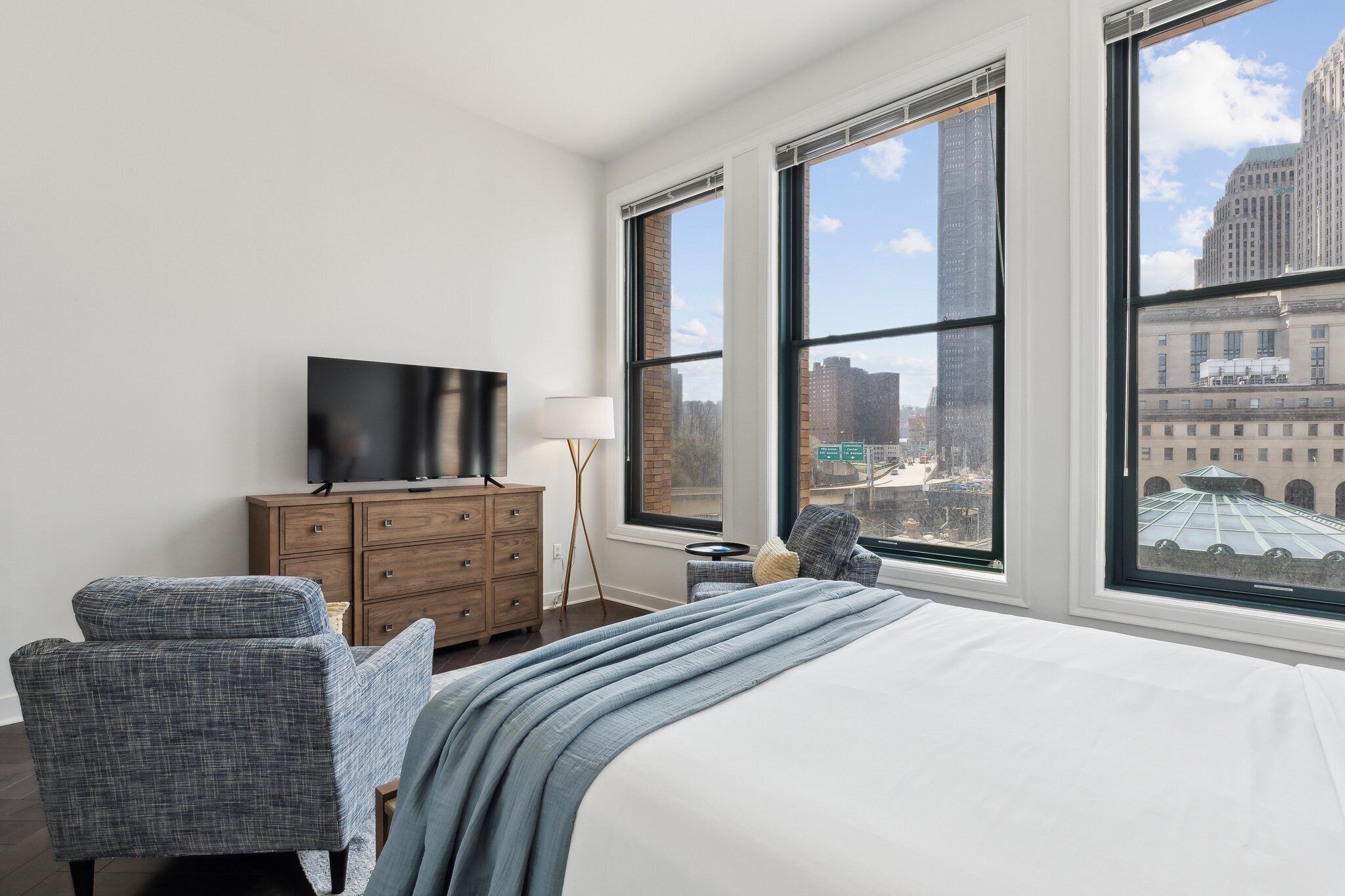 A hotel room with a large bed, a TV on a wooden dresser, a chair, and a view of city buildings through large windows.