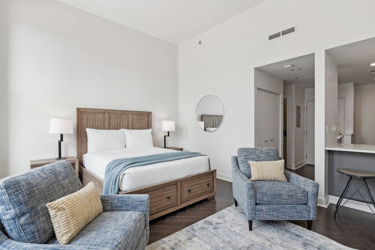 Un dormitorio moderno con una cama de madera, dos sillones, un escritorio pequeño y un espejo redondo en la pared.