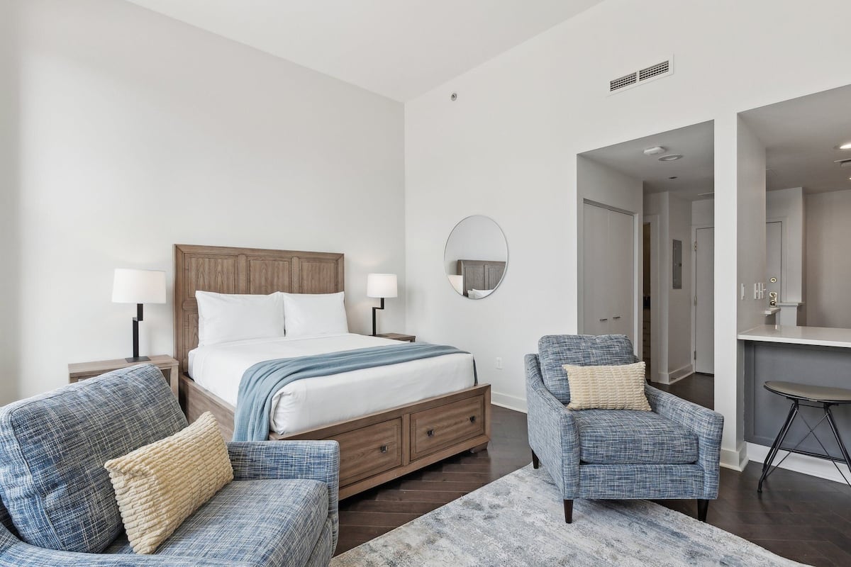 A modern bedroom with a wooden bed, two armchairs, a small desk, and a round mirror on the wall.