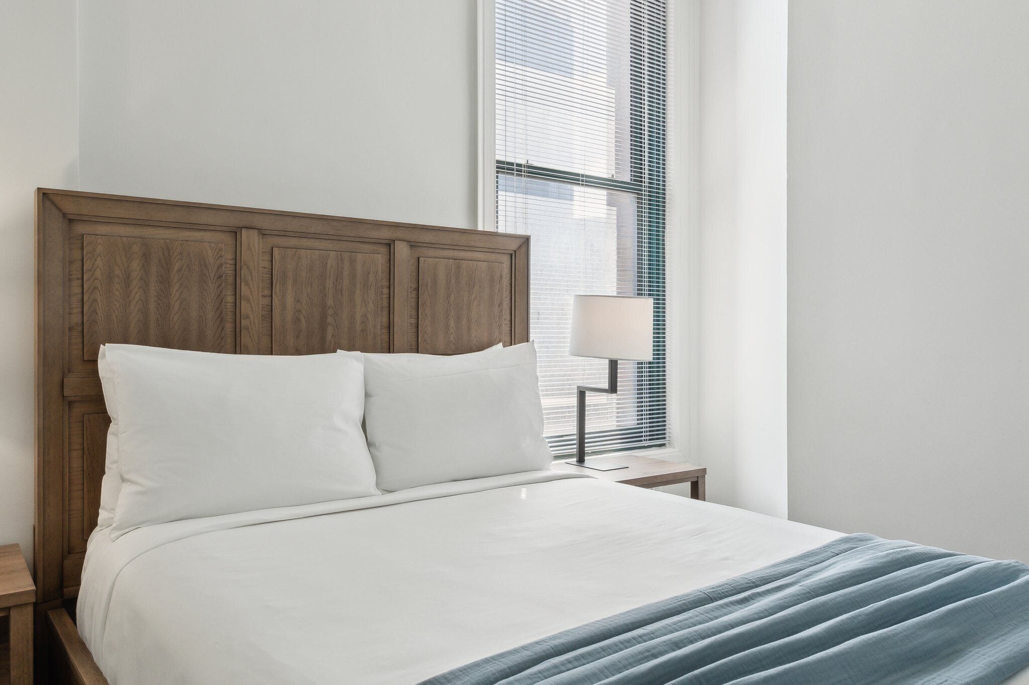 Una cama bien tendida con cabecera de madera, una lámpara de noche y una ventana con persianas.