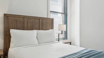 Una cama bien tendida con cabecera de madera, una lámpara de noche y una ventana con persianas.