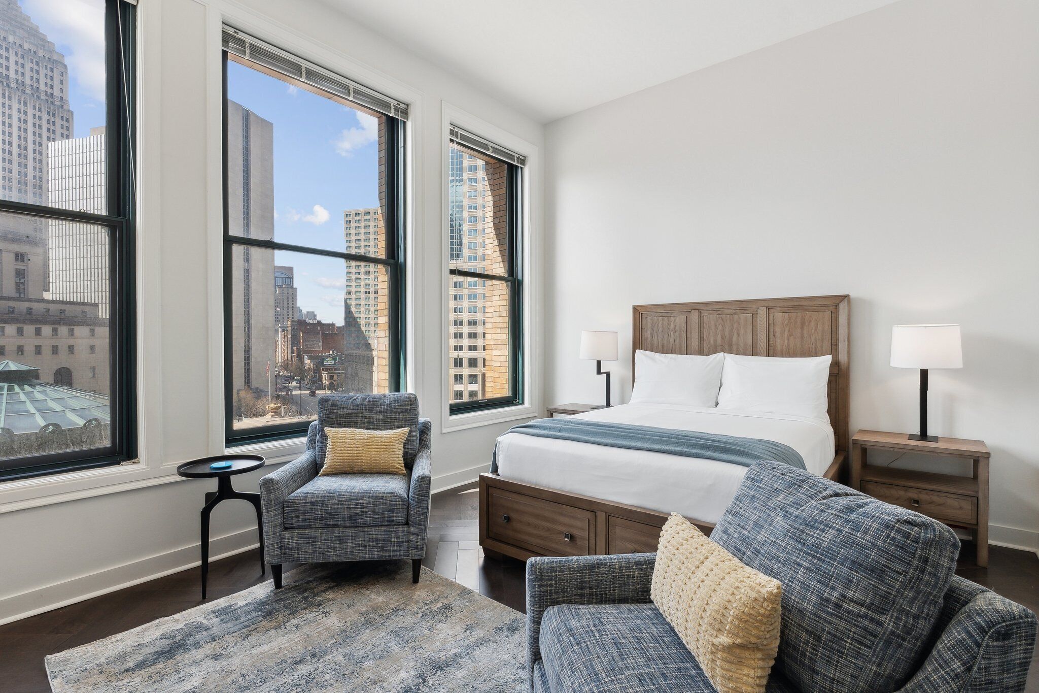 Ein Hotelzimmer mit einem großen Bett, zwei Sesseln, einem kleinen Tisch und Blick auf die Stadt durch große Fenster.
