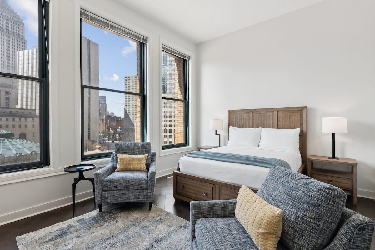 Habitación de hotel con una cama grande, dos sillones, una mesita y vistas a la ciudad a través de amplios ventanales.