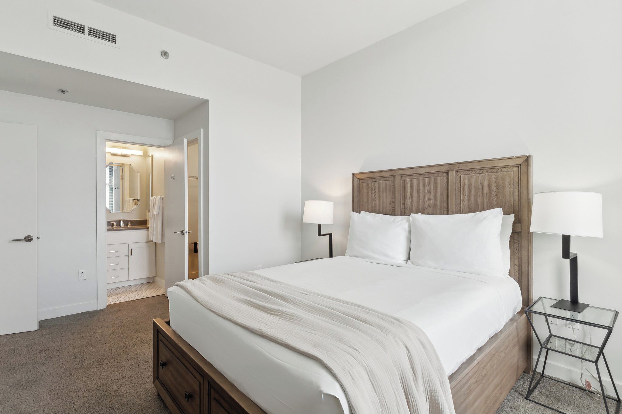 A neatly arranged bedroom with a large bed, bedside lamps, and a visible bathroom in the background.