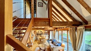 Amplio espacio tipo loft con techo alto, vigas de madera expuestas, una escalera y grandes ventanales que ofrecen vistas al exterior.