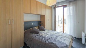 A bedroom with a bed, wooden wardrobe, and a view of the city through a large window. - Apartment in Oostduinkerke Near the Coast (Koksijde)