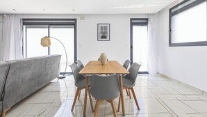 A modern dining area with a wooden table and grey chairs, large windows with curtains, and a grey sofa. - Appartement de 3 pieces a Birkhadem (Algiers)