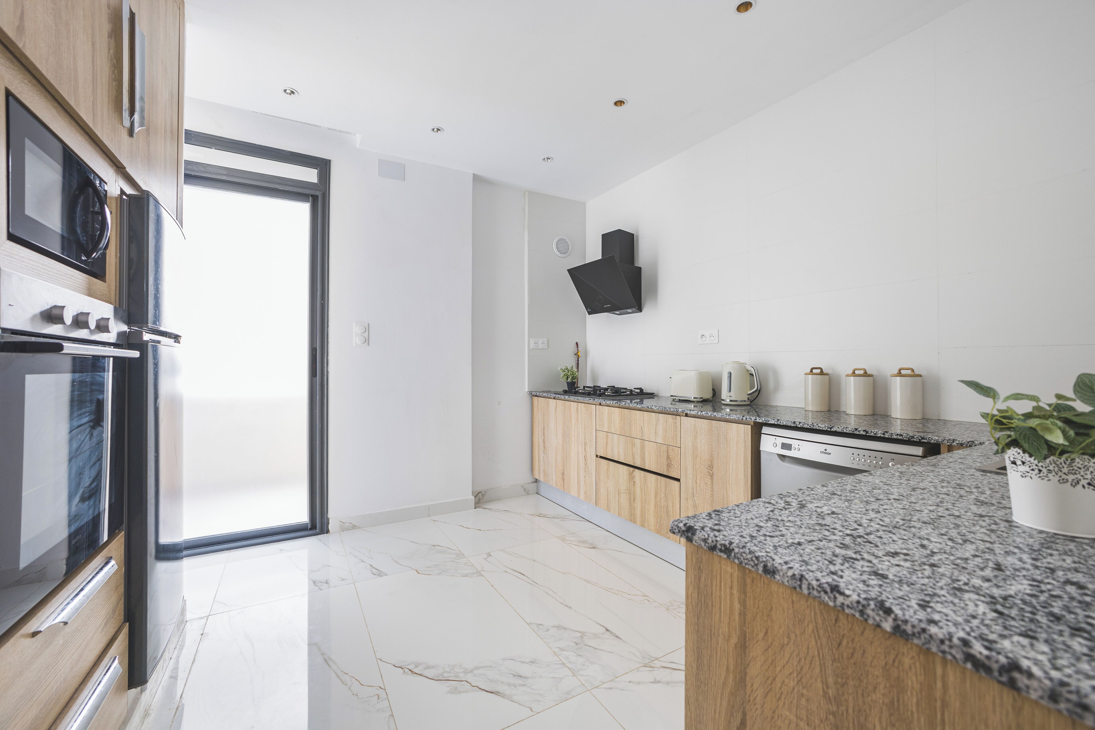 Cozinha moderna com forno embutido, micro-ondas e máquina de lavar louça. Há uma grande janela que proporciona luz natural e vista para o exterior.