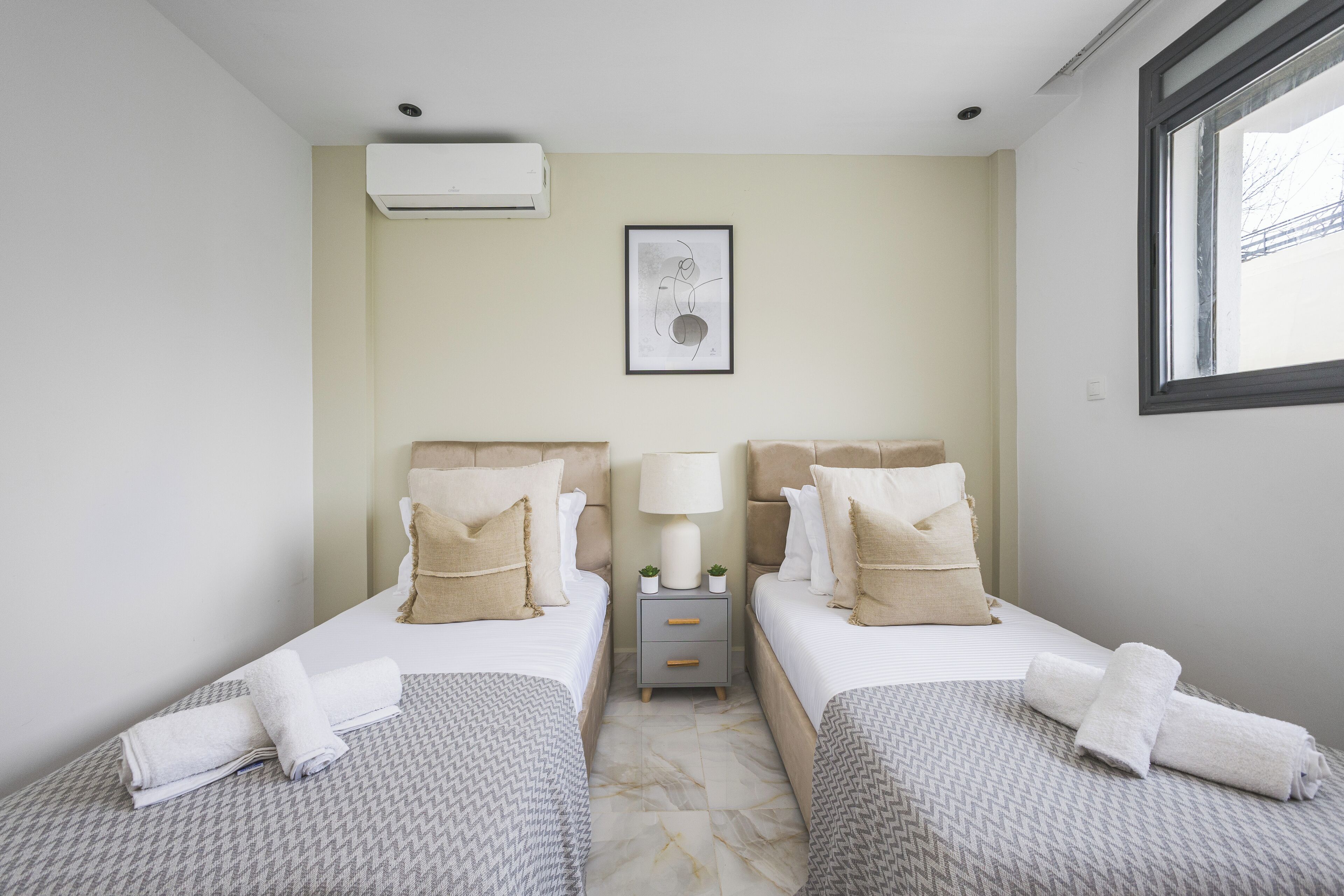 Hotelkamer met twee bedden, een nachtkastje met een lamp, een ingelijst schilderij aan de muur en een airconditioner.