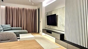 A modern living room with a sectional sofa, a flat-screen TV mounted on a wall, and a wooden floor. - Air Onia (Budva)