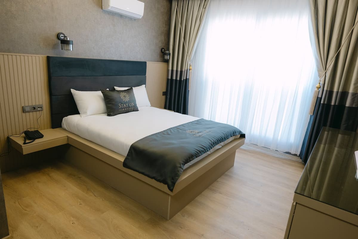 A modern hotel room with a large bed, bedside tables, and a view of the outdoors through sheer curtains.