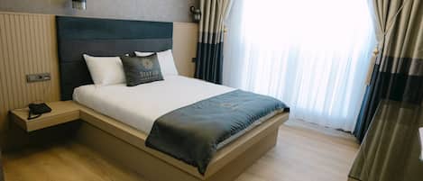 A modern hotel room with a large bed, bedside tables, and a view of the outdoors through sheer curtains.
