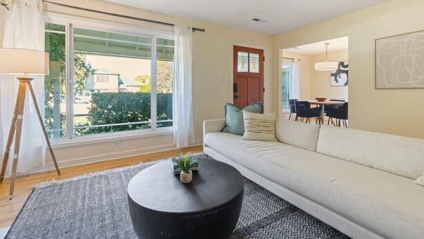 A modern living room with a large window, a black round coffee table, a beige sofa, and a dining area in the background. - @ Marbella Lane - Modern and Sleek Home in Redwood (Redwood City)