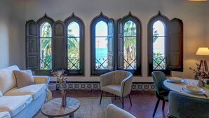 Una habitación con sofá, sillas, una mesita y una mesa de comedor con sillas. Amplias ventanas ofrecen vistas al mar y a las palmeras.