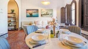 Un comedor con una mesa redonda dispuesta para cuatro personas, rodeada de sillas. Hay dos cuadros en la pared, una planta en maceta a la izquierda y una estantería.
