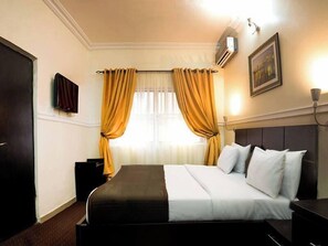 Habitación de hotel con cama, mesita de noche, televisor, ventana con cortinas y equipo de aire acondicionado.