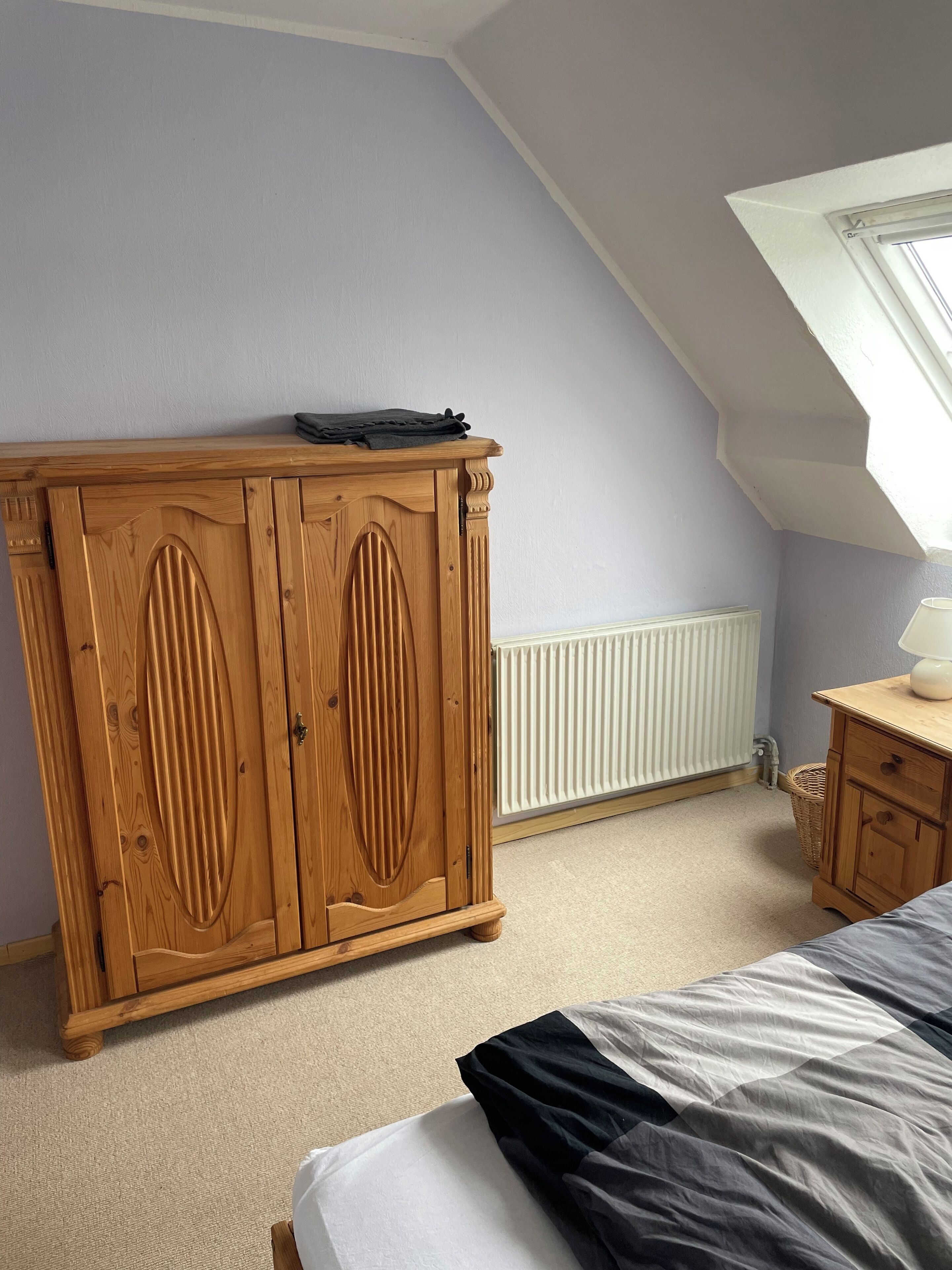A bedroom with a sloped ceiling, a wooden wardrobe, a bedside table with a lamp, and a radiator.