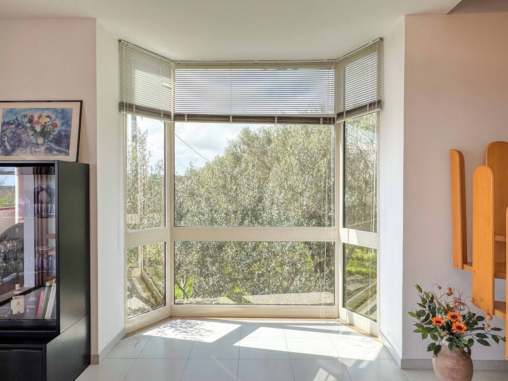 Ein Eckzimmer mit großem Fenster, einem gerahmten Bild an der Wand und einer Vase mit Blumen.