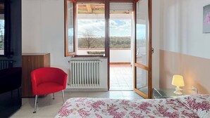 Un dormitorio con una cama, una silla roja, un radiador y vistas a través de un ventanal.