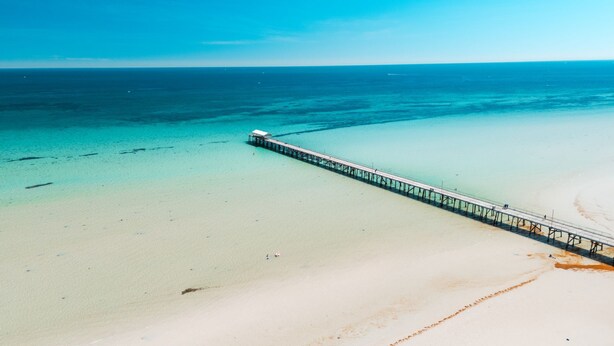 A long pier extending into a clear blue sea from a sandy beach.