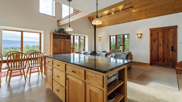 A spacious kitchen with wooden cabinets, a large island, and pendant lighting. There are large windows offering views of the outdoors.