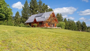A log cabin with a sloped roof, surrounded by a grassy field and dense trees.