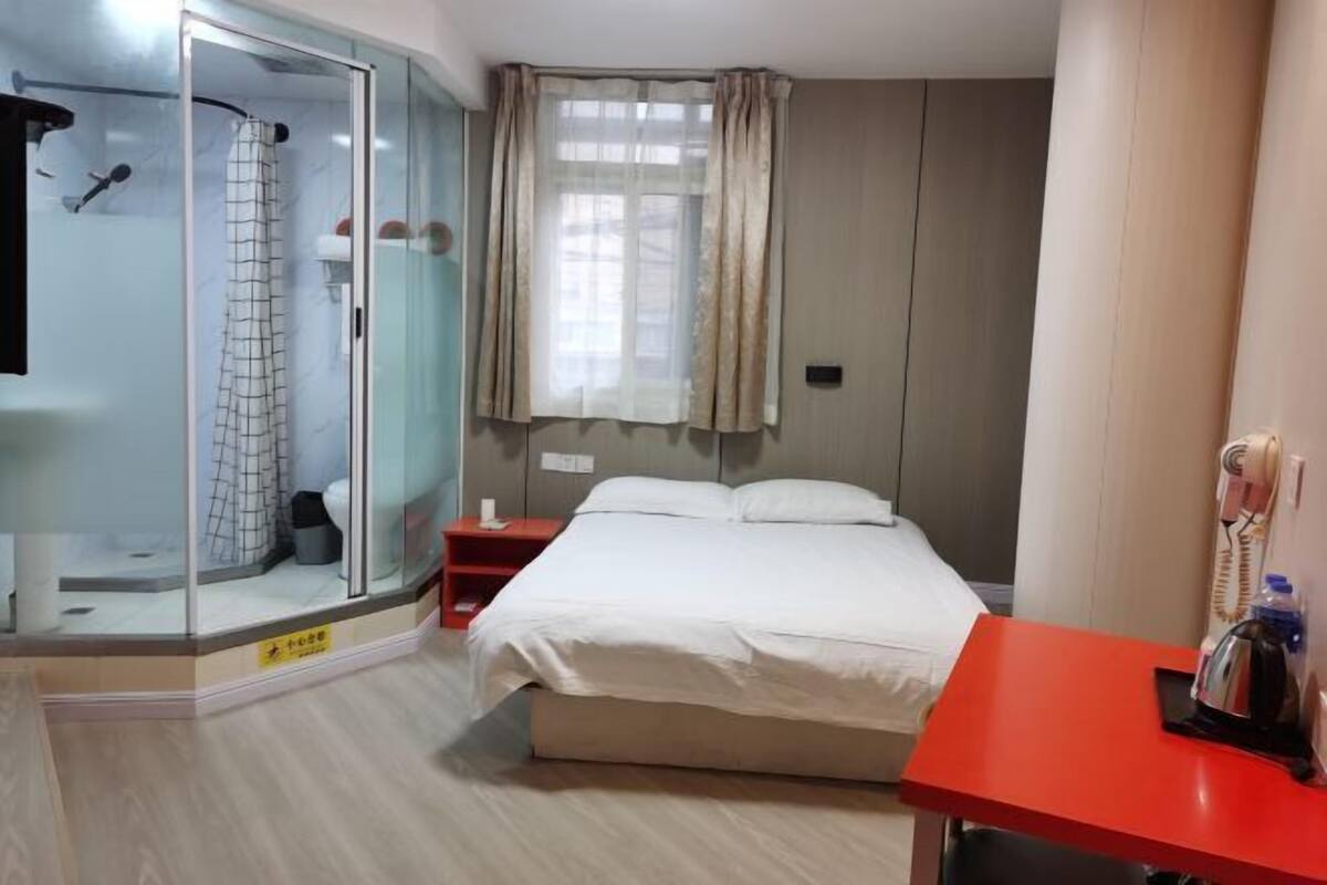 A hotel room with a glass shower enclosure, a bed with white bedding, a red nightstand, and a red desk with a kettle.