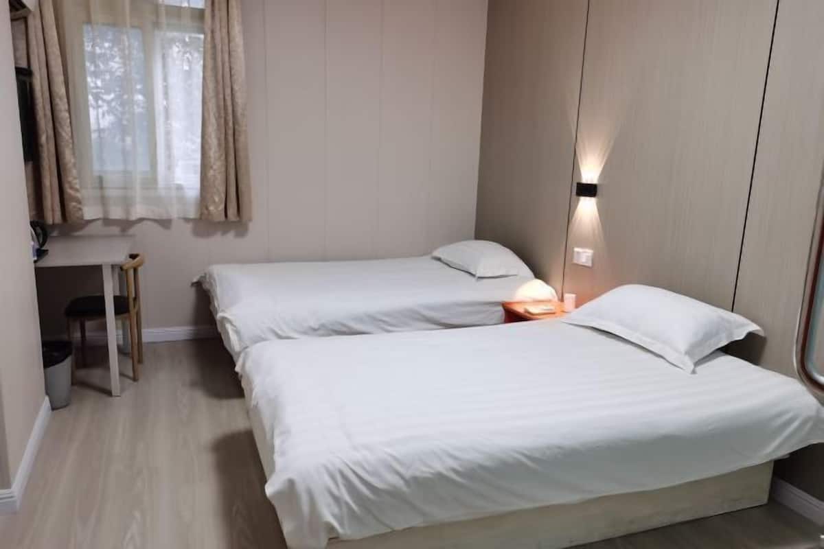 A small, clean room with two single beds, a window with curtains, a bedside table with a lamp, and a chair.