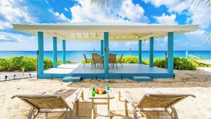 A beachside outdoor seating area with a covered stage, chairs, and a table with drinks and fruit.