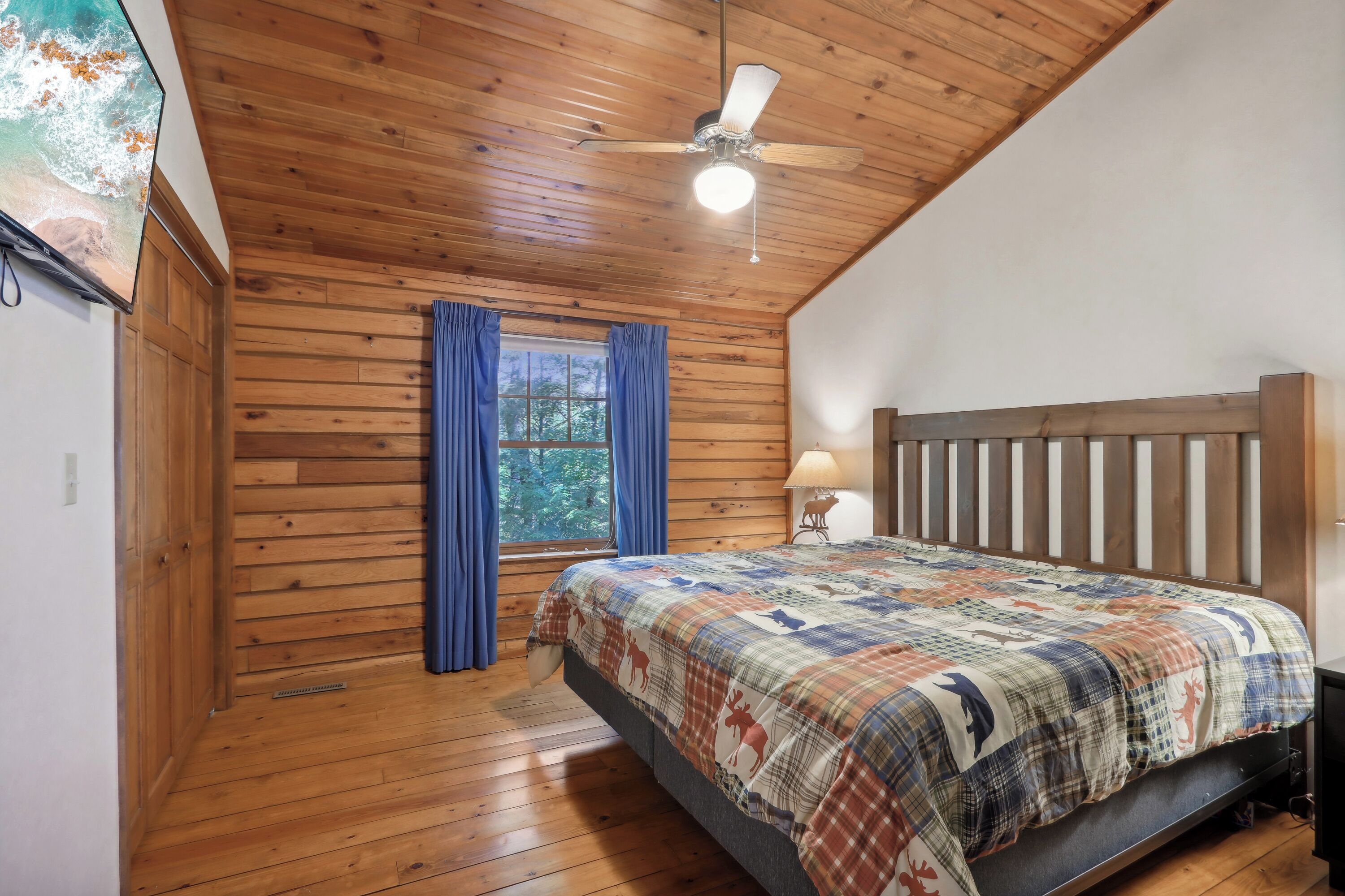 Un dormitorio con paneles de madera, una cama, un ventilador de techo, una ventana con cortinas y una lámpara.