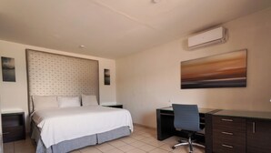 Une chambre d’hôtel avec un lit, un bureau et une chaise. Il y a un tableau accroché au mur et un appareil de climatisation au-dessus du bureau.