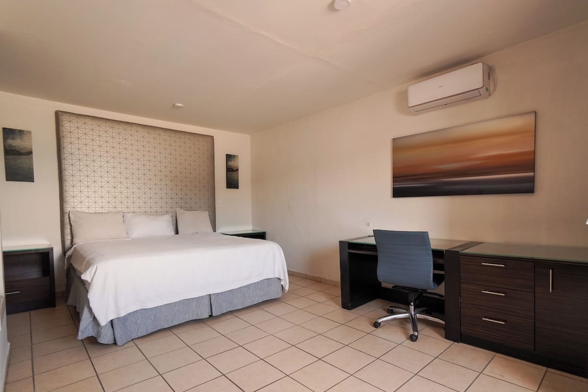 A hotel room with a bed, a desk, and a chair. There is a painting on the wall and an air conditioning unit above the desk.
