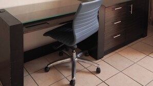 Un espace de bureau moderne avec un bureau en verre, un fauteuil de bureau noir, un meuble en bois foncé doté de tiroirs et un climatiseur fixé au mur.