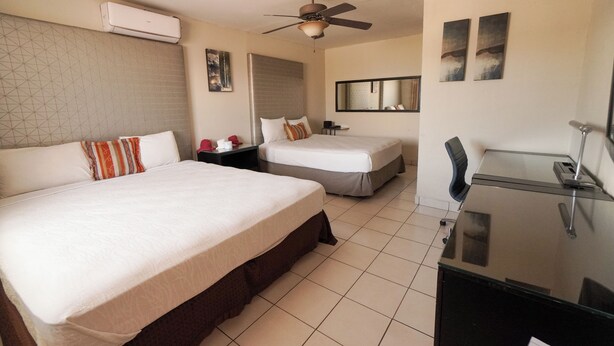 Une chambre d’hôtel avec deux lits, un bureau, une chaise, un ventilateur de plafond et un grand miroir.
