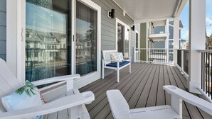Une terrasse en bois couverte, agrémentée de meubles d’extérieur blancs, notamment d’un banc et de chaises, et de grandes fenêtres avec des rideaux.