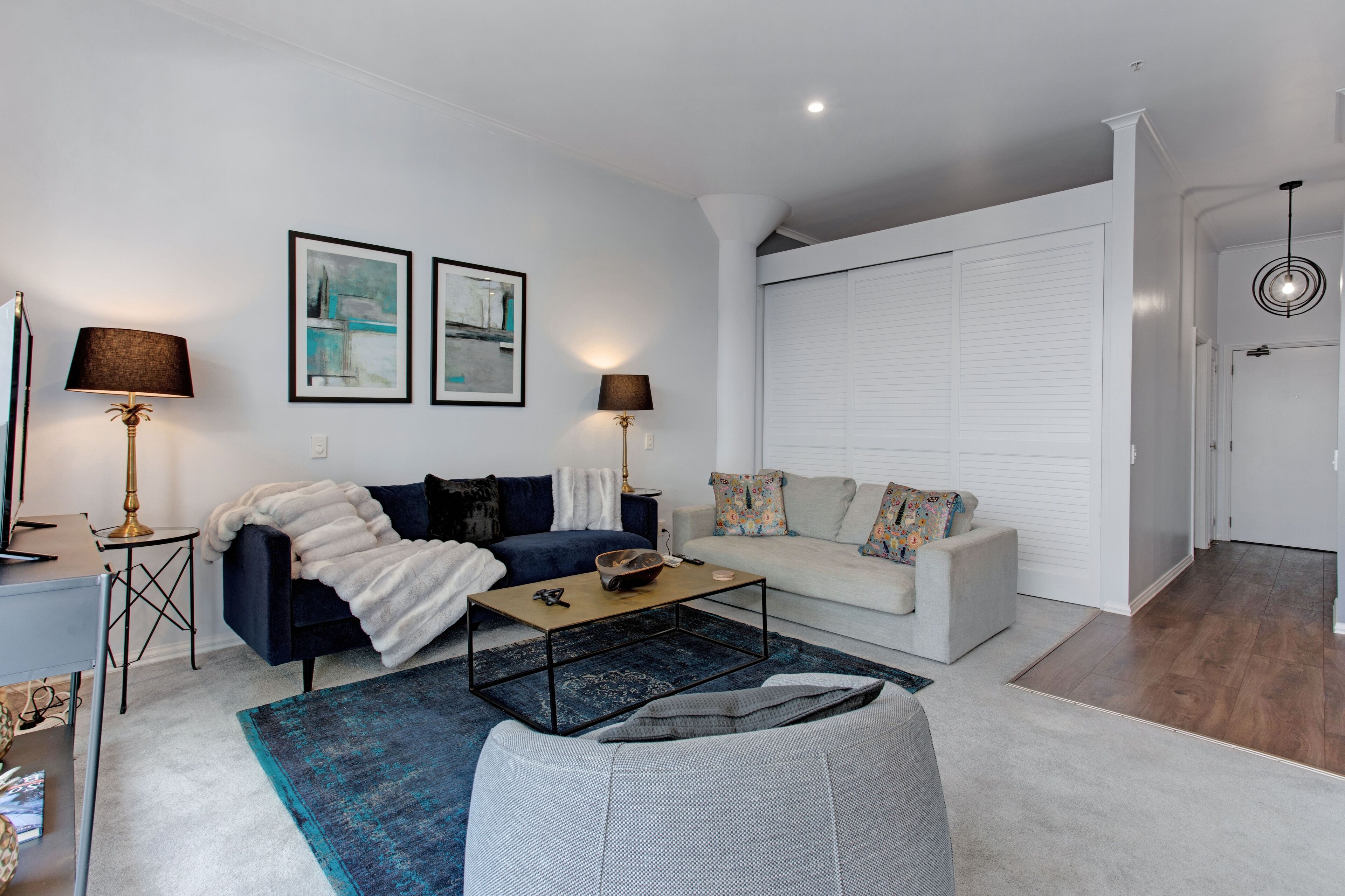 A modern living room with a dark blue sofa, a white sofa, a coffee table, and a blue rug. There are two lamps, a framed artwork on the wall, and a white door in the background.