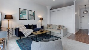 Una sala moderna con un sofá azul oscuro, un sofá blanco, una mesa de centro y un tapete azul. Se observan dos lámparas, una obra de arte enmarcada en la pared y una puerta blanca al fondo.