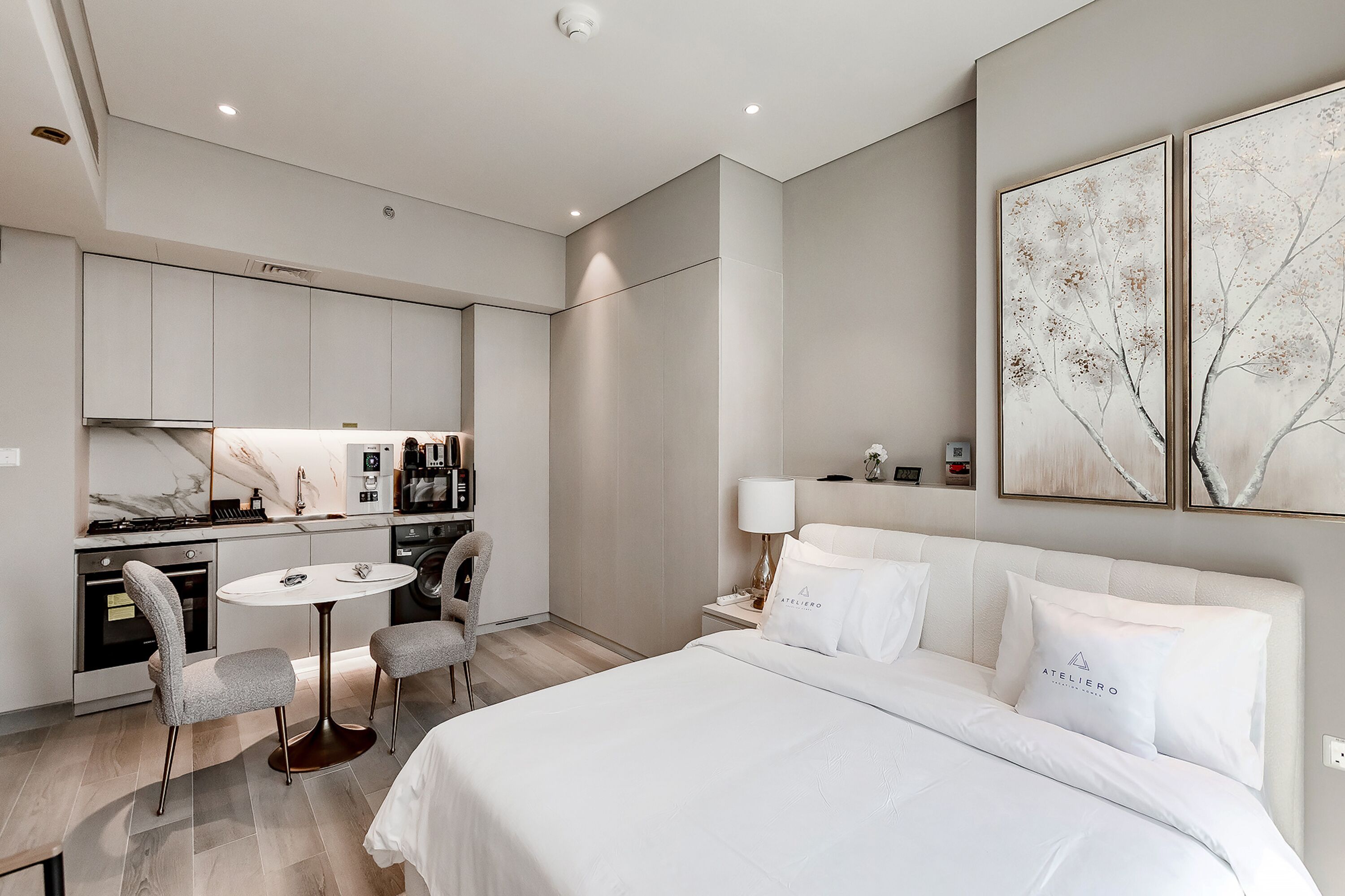 Une chambre d’hôtel moderne dotée d’un grand lit, d’une table à manger avec des chaises, d’un petit coin cuisine et d’œuvres d’art accrochées aux murs.