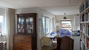 A room with a wooden cabinet, a dining table with chairs, a bookshelf, and a window with a view. - Apartment With Dunes View of Petten (Petten)