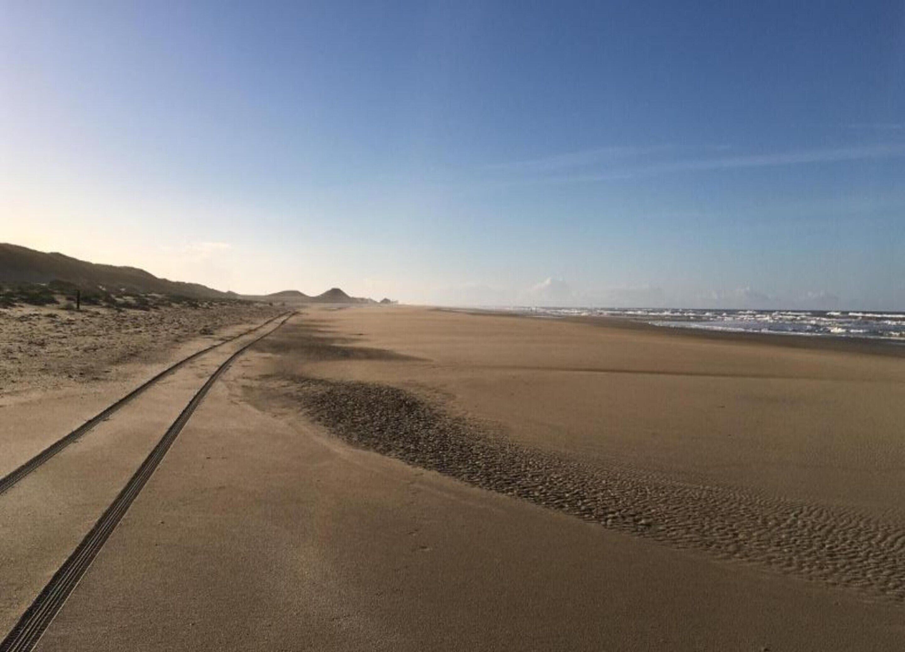 A wide, empty beach with a clear sky and distant mountains.