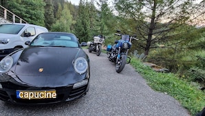 Une voiture noire, avec une plaque d’immatriculation bien visible, est garée sur un chemin pavé, avec un mur de pierres et des arbres en arrière-plan.