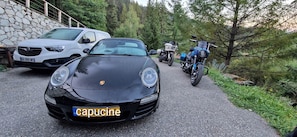 Un coche negro con una placa de matrícula visible, estacionado en un camino pavimentado con un muro de piedra y árboles de fondo.