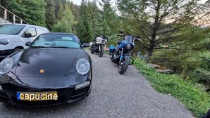 Un coche negro con una placa de matrícula visible, estacionado en un camino pavimentado con un muro de piedra y árboles de fondo.