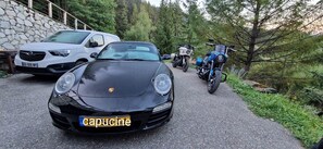 Un coche negro con una placa de matrícula visible, estacionado en un camino pavimentado con un muro de piedra y árboles de fondo.