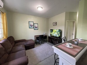 Ein Wohnzimmer mit braunem Sofa, einem Fernseher, einem Schreibtisch mit Stuhl und einem Esstisch mit Stühlen.