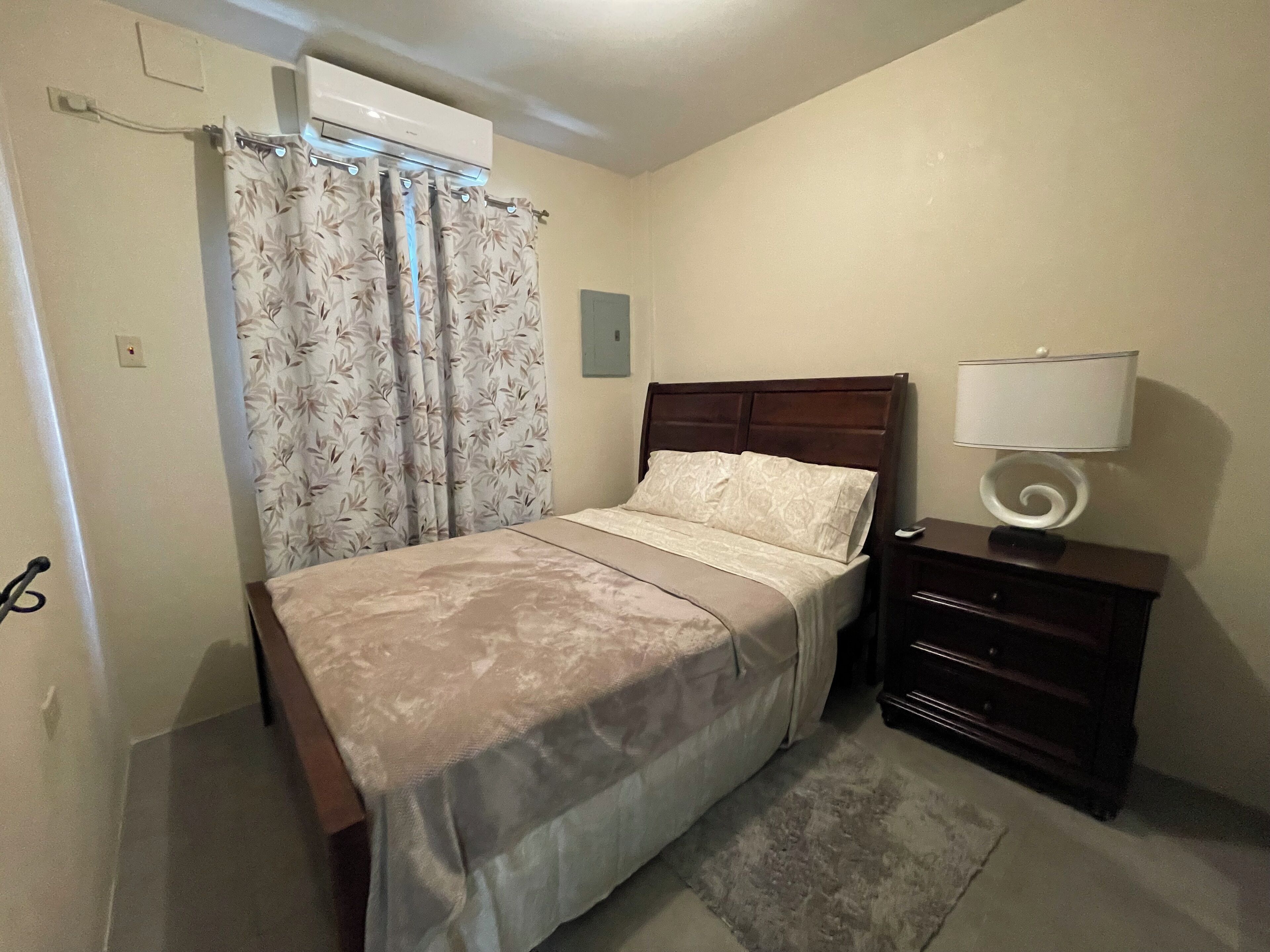 A bedroom with a bed, a nightstand with a lamp, a wooden headboard, and a window with curtains.