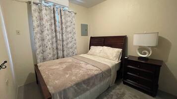 A bedroom with a bed, a nightstand with a lamp, a wooden headboard, and a window with curtains.