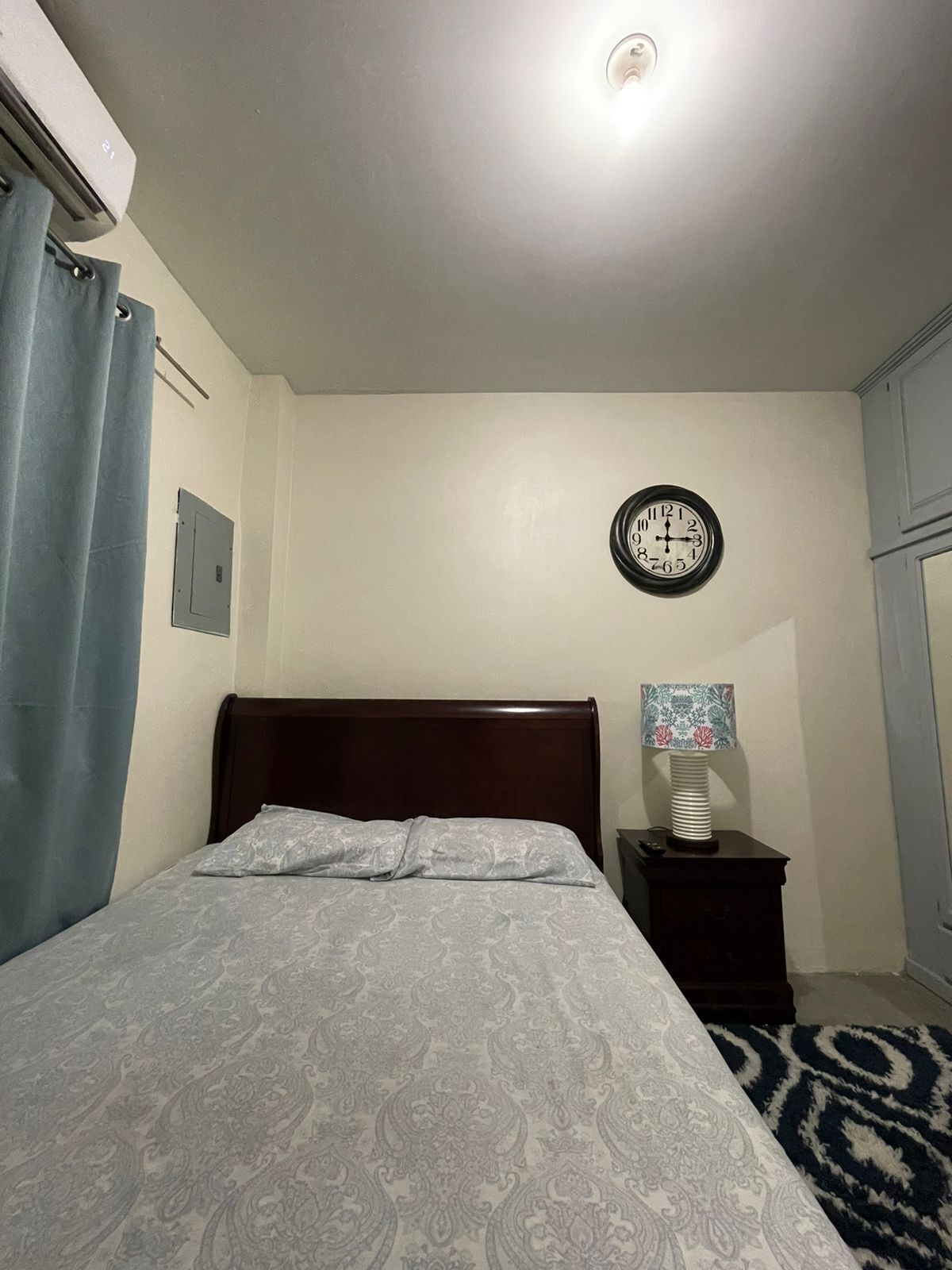 A bedroom with a bed, a wall clock, a bedside lamp, and a window with curtains.
